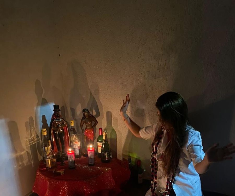 Mãe Stefany de jaleco branco e guias, com as mãos erguidas diante do altar com imagens, velas vermelhas e toalha vermelha.
