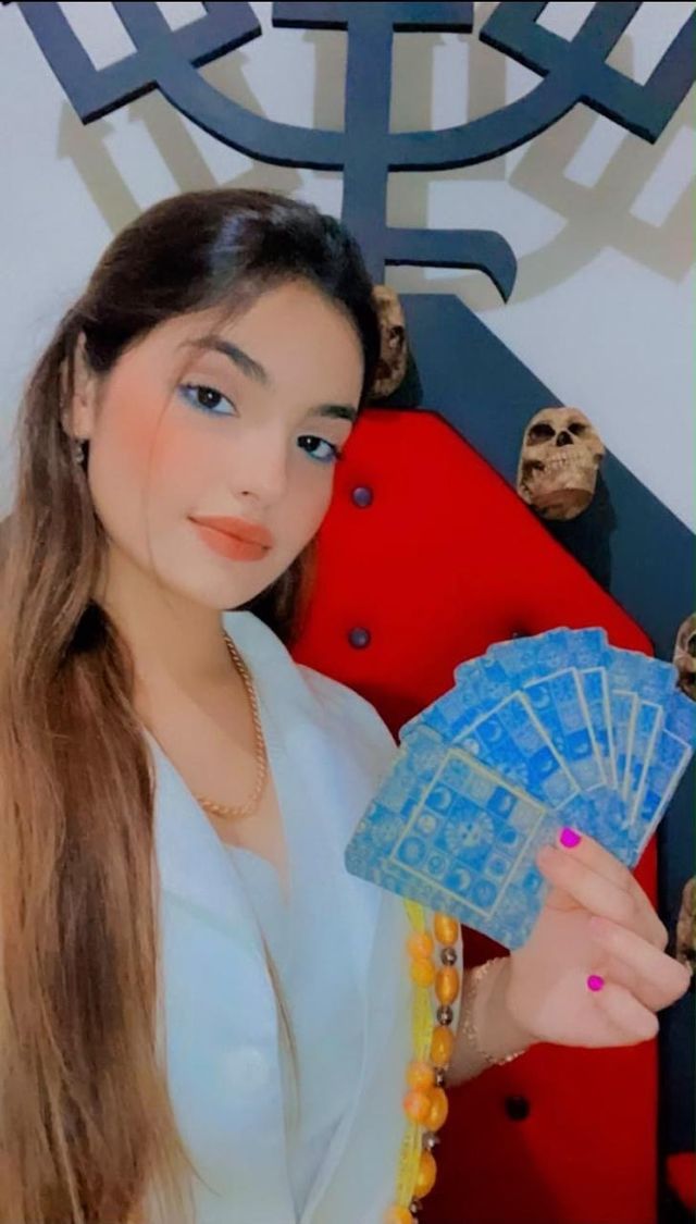 Mãe Stefany segurando um leque de cartas azuis, de roupa branca e guias de contas laranja.