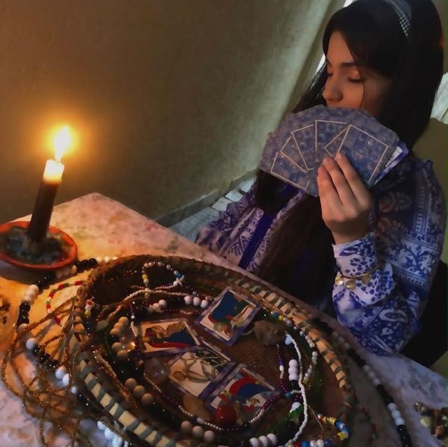 Mesa de consulta com vela acesa, guias de contas e cartas abertas sobre uma cesta, enquanto Mãe Stefany segura o baralho.